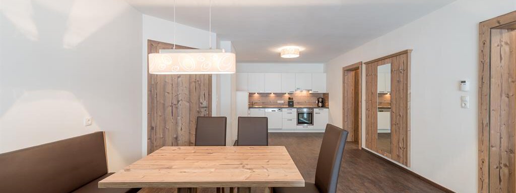 A modern dining room with a large wooden table and a cozy bench. In the background, an open kitchen can be seen. - Revmatologická poradna Appt Top 4 - Sölden