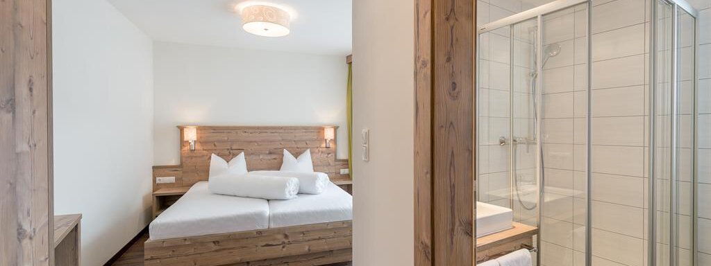 A modern room with a wooden double bed and two pillows. In the background, there is a bathroom with a modern shower cubicle. - Revmatologická poradna Appt Top 4 - Sölden