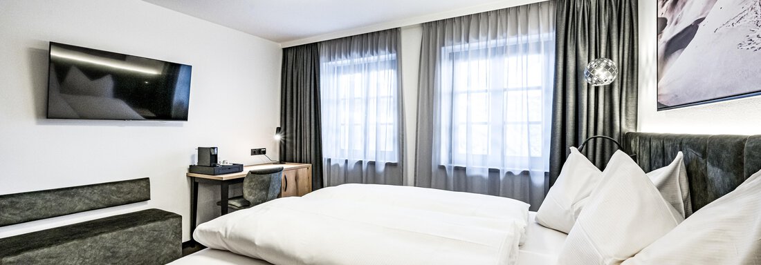 A modern hotel room with a large bed and two pillows. There is a television, a desk, and large windows with curtains. - Sonnalp Residences - Sölden