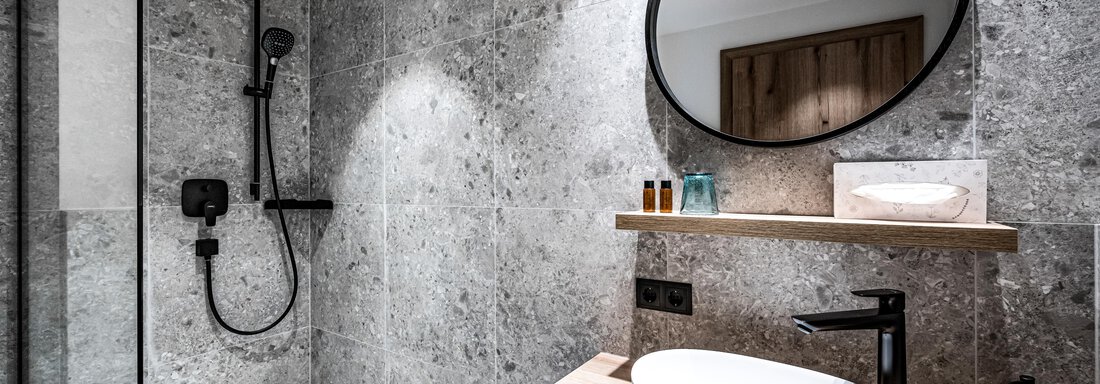 A modern bathroom with a walk-in shower and an elegant sleek sink unit. The walls are clad in grey stone, and there is a round mirror above the sink. - Sonnalp Residences - Sölden