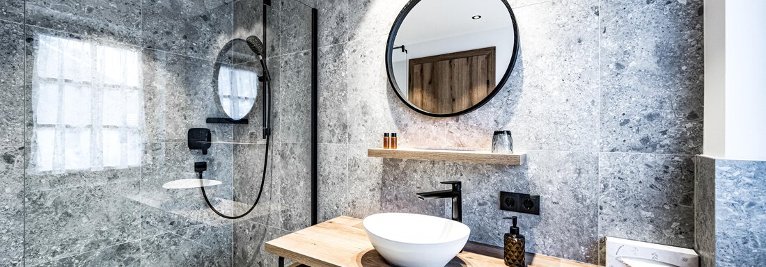 A modern bathroom with gray stone walls and an elegant shower. A sink is placed on the wooden shelf, and decorative elements add a stylish accent. - Sonnalp Residences - Sölden