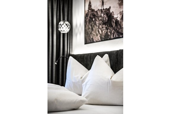 A modern bedroom with white pillows on a dark headboard. In the background, you can see an artistic wall hanging and a stylish lamp. - Sonnalp Residences - Sölden
