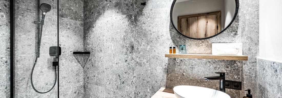 A modern bathroom with an elegant shower and a stylish sink ensemble. The walls are designed with gray tiles and there is a round mirror surface. - Sonnalp Residences - Sölden