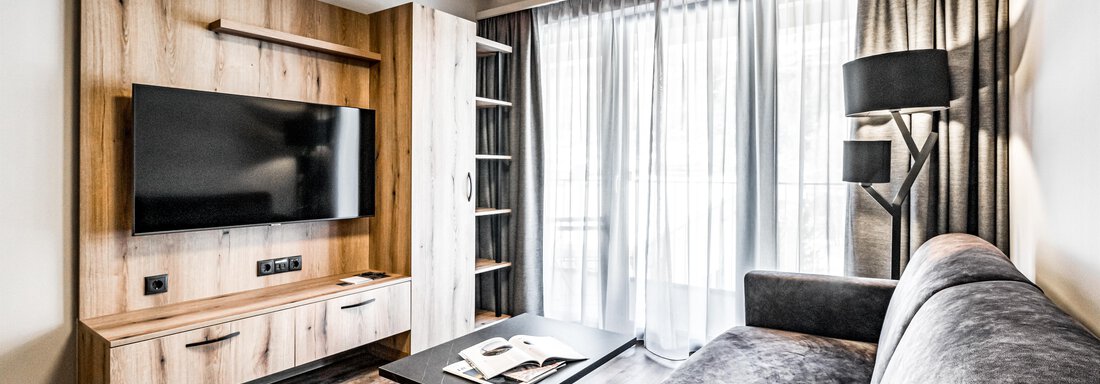 A modern living room with a gray sofa and a large TV on the wall. Large windows provide plenty of natural light. - Sonnalp Residences - Sölden