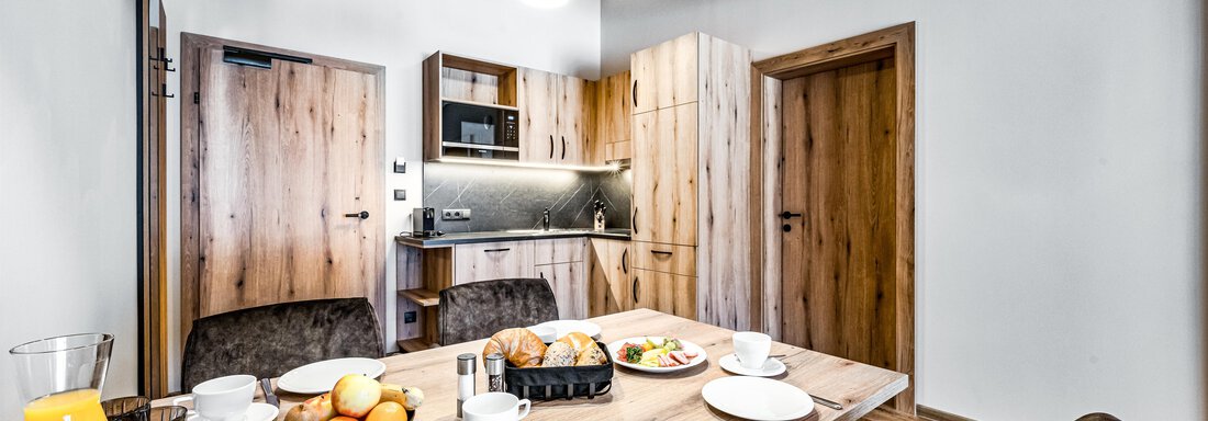 A modern furnished dining area with a wooden table, chairs, and a set table. In the background, there is a kitchen corner with wooden shelves and modern lights. - Sonnalp Residences - Sölden
