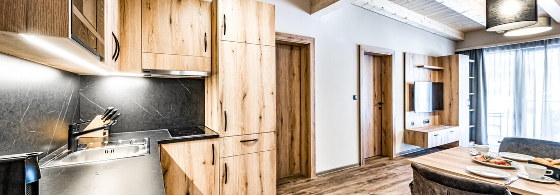 A modern kitchen with wooden cabinets and a black countertop. The room is bright and stylishly furnished, with a table and chairs in the background. - Sonnalp Residences - Sölden