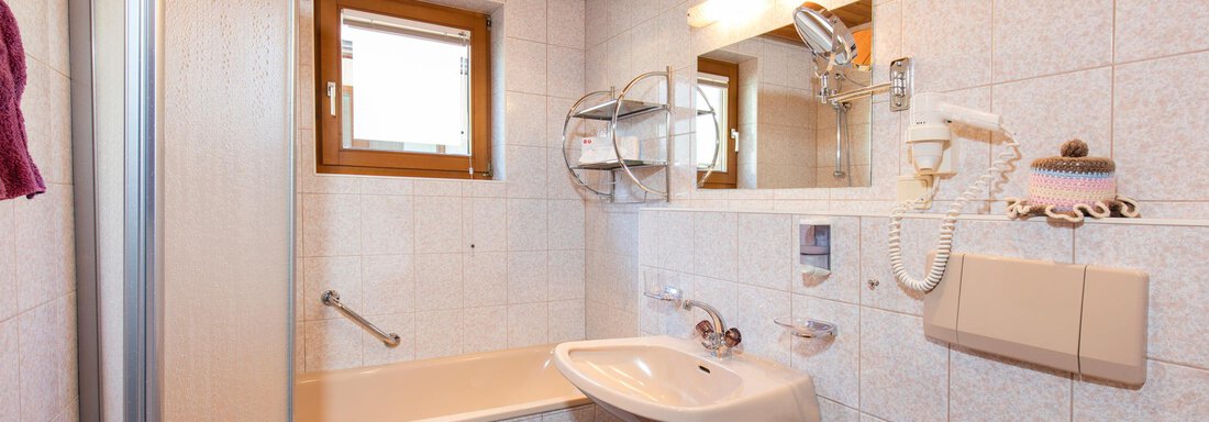A modern bathroom with a bathtub and a sink. The walls are tiled, and there is a window as well as mirrors. - Sportpension Rubin - Sölden