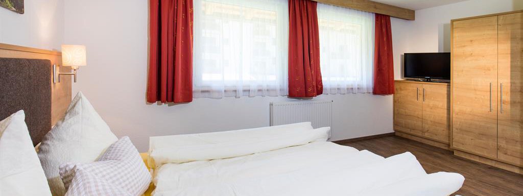 A bright bedroom with a large bed and red curtains. There is a wardrobe and a television in the room. - Sportpension Rubin - Sölden