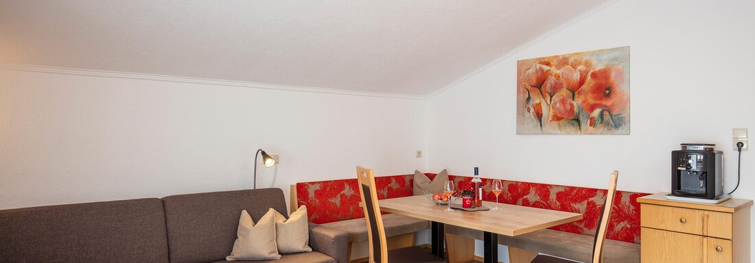 A cozy living space with a beige couch and a dining table. A picture with red floral motifs hangs on the wall. - Sportpension Rubin - Sölden