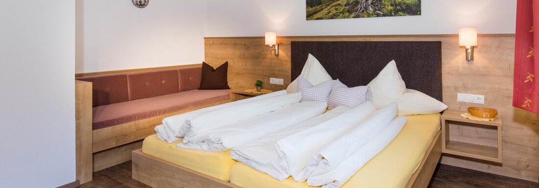 A cozy bedroom with a large double bed and bright, warm colors. On the wall hangs a piece of artwork depicting a landscape, and there is a seating area with a bench. - Sportpension Rubin - Sölden
