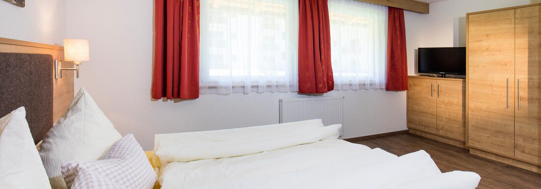 A cozy bedroom with a double bed and large windows. The red curtains and wooden furniture give the room a warm atmosphere. - Sportpension Rubin - Sölden