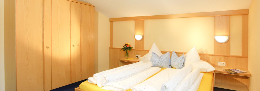 A cozy bedroom with a double bed and yellow bedcovers. The room is bright and has a simple wooden wardrobe as well as nice decoration. - Sportpension Rubin - Sölden