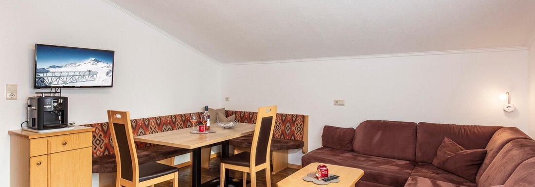 wohnhaft A cozy living space with a brown sofa, a dining table, and four chairs. A television is mounted on the wall and the floor is made of wood. - Sportpension Rubin - Sölden