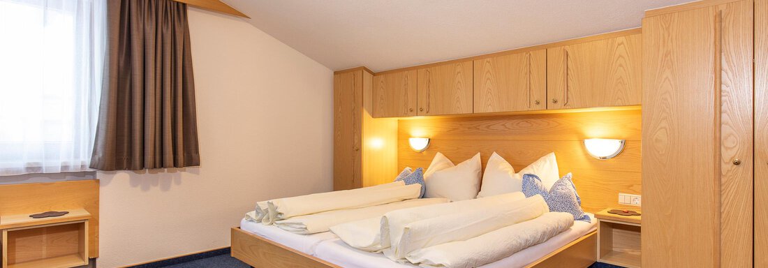 A quiet bedroom with a large bed and wooden wardrobes. The room is bright and friendly with carpet flooring and a window with curtains. - Sportpension Rubin - Sölden