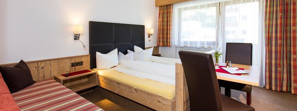 A modern hotel room with a large window and plenty of natural light. It features a double bed, a seating area, and a desk. - Sportpension Rubin - Sölden