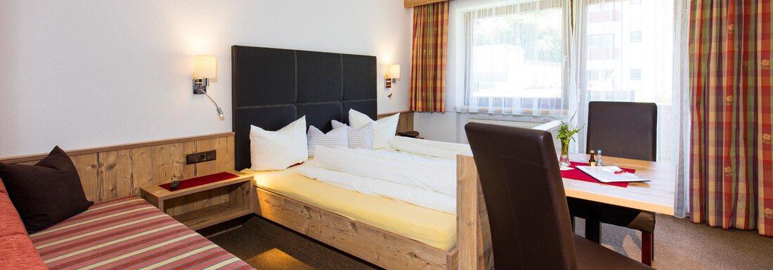 A bright room with a large window and cozy furnishings. There is a double bed, a desk, and a seating area. - Sportpension Rubin - Sölden