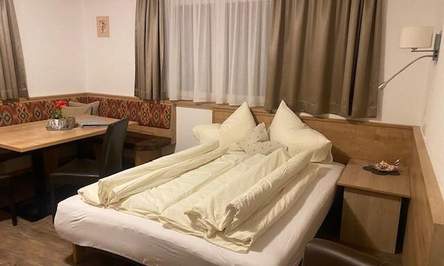 A cozy room with a large bed and two pillows. There is a small table and chairs, as well as decorative window curtains. - Sportpension Rubin - Sölden