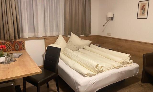 A cozy bedroom with a double bed and a wooden table. The walls are painted in warm tones and there is a pleasant window decoration. - Sportpension Rubin - Sölden