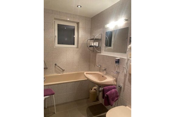 A modern bathroom with a bathtub and a sink. The walls are tiled and there is a mirror as well as bright lighting. - Sportpension Rubin - Sölden