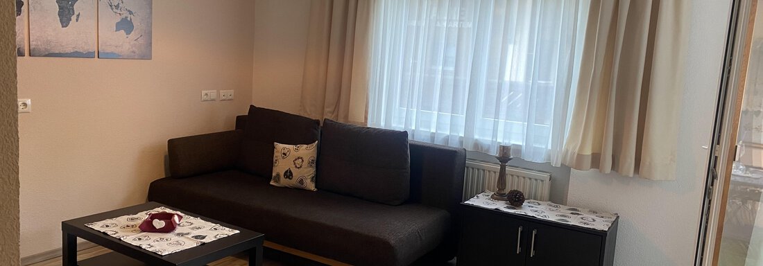 A modern living room with a dark sofa and a coffee table. Large windows let in plenty of light and there are decorative wall pictures. - Sportpension Rubin - Sölden