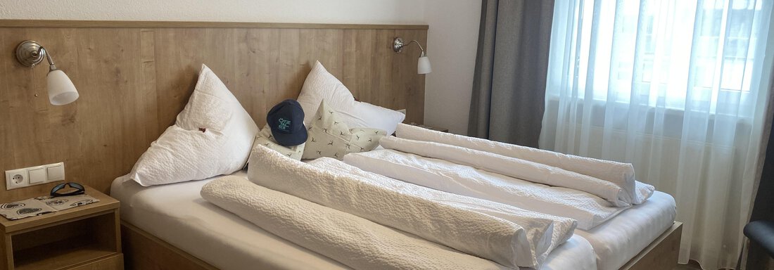 A bright bedroom with a double bed and two pillows. The windows are equipped with bright curtains and there is a bedside table next to it. - Sportpension Rubin - Sölden