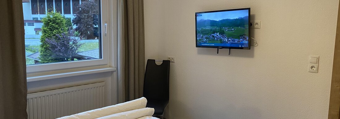 A cozy bedroom with a bed and a TV on the wall. Through the window, one can see a green landscape. - Sportpension Rubin - Sölden