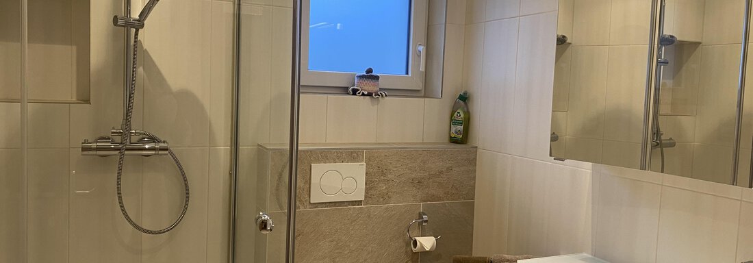 A modern bathroom with a shower, toilet, and a sink. The walls are light tiled, and a window provides natural light. - Sportpension Rubin - Sölden