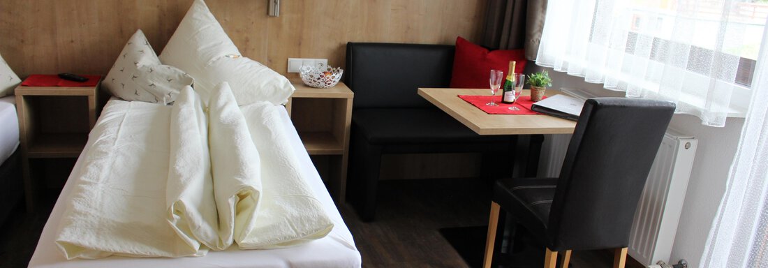 A cozy room with two beds and a small dining area. The room has large windows with daylight and warm wooden paneling. - Sportpension Rubin - Sölden