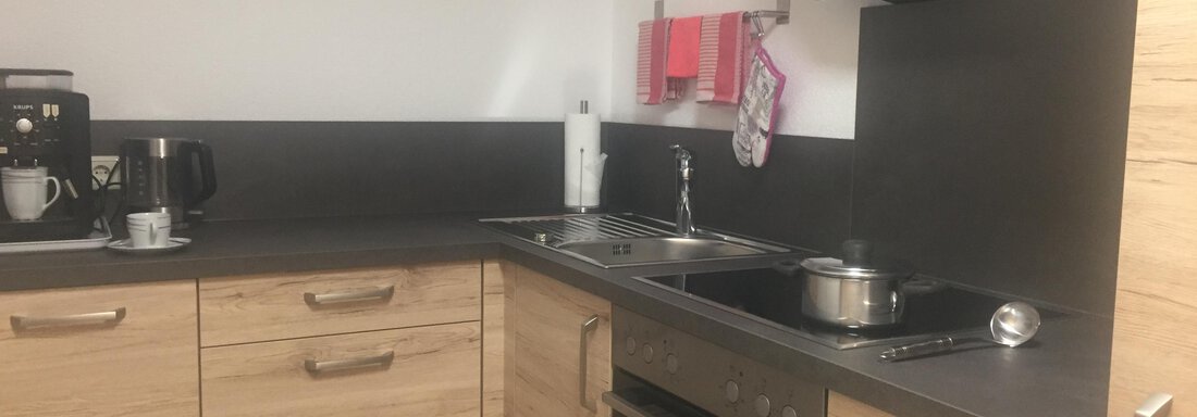 A modern kitchen with wooden cabinets and a dark countertop. Various kitchen appliances are visible, such as a coffee maker and a stove. - Sportpension Rubin - Sölden