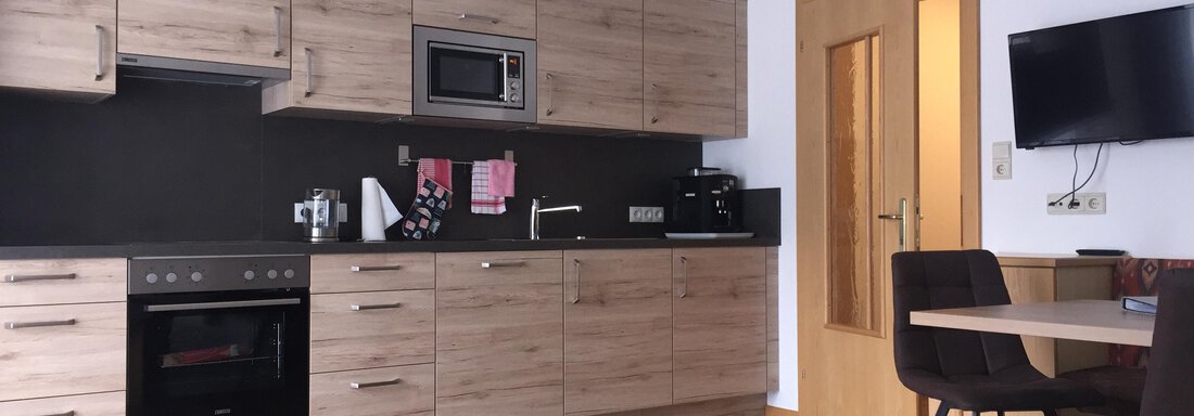 A modern kitchen with wooden cabinets and a dining area. A television hangs on the wall and the floor is made of wood. - Sportpension Rubin - Sölden