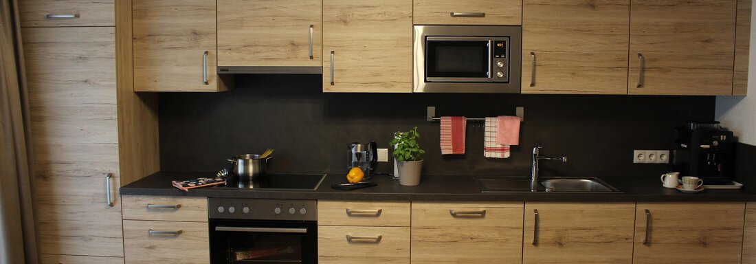 A modern kitchen with wooden cabinets and a dark countertop. On the countertop, there are some kitchen utensils and plants. - Sportpension Rubin - Sölden