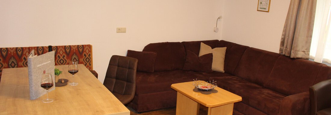 A cozy living room with a brown couch and a small table. The dining area consists of a wooden table and chairs, decorated with a bottle of wine. - Sportpension Rubin - Sölden