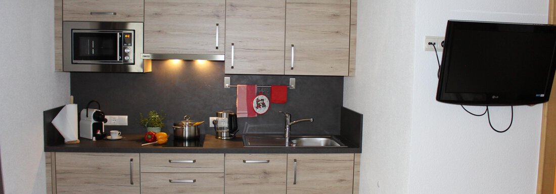 IMG_3687 A modern kitchen with wooden cabinets and a countertop made of dark material. There is a microwave, a stove, and a sink. - Sportpension Rubin - Sölden