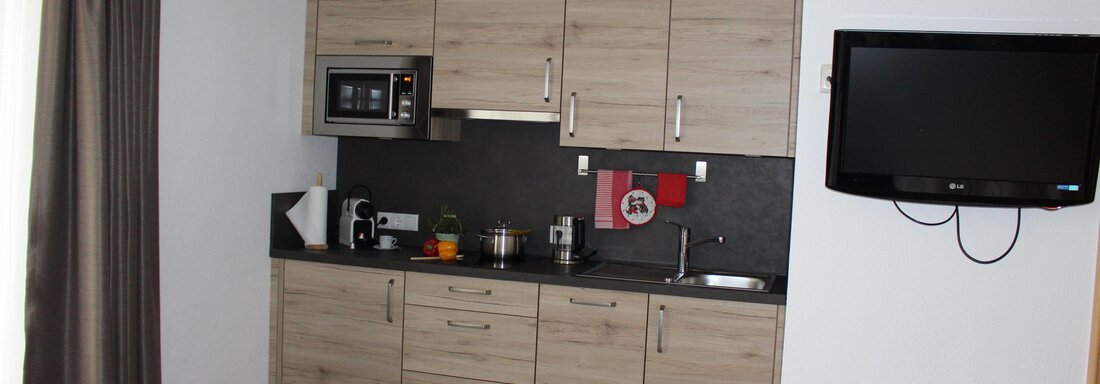 A modern kitchen with wooden surfaces and a cooking area. A TV is mounted on the wall, and there are curtains next to a window. - Sportpension Rubin - Sölden