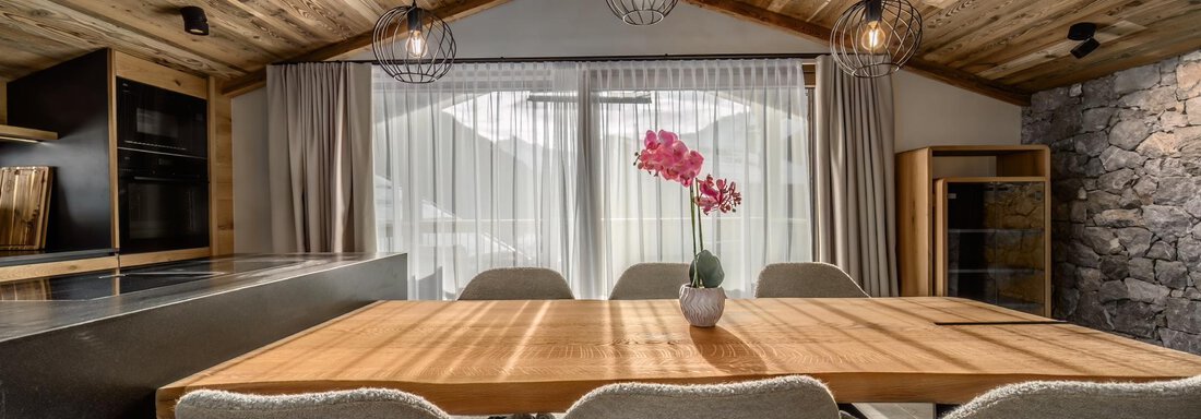 A modern dining room with a long wooden table and comfortable chairs.  
Large windows let in plenty of light, and an orchid adds a touch of color to the space. - The Peak Sölden - Sölden