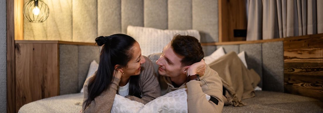 A couple lovingly smiles at each other as they lie on a round bed. The cozy environment with warm light creates an intimate atmosphere. - The Peak Sölden - Sölden