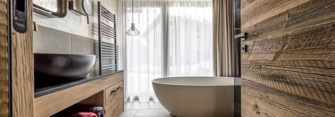 A modern bathroom with a freestanding tub, wooden and stone accents. Light streams through large windows, creating an inviting atmosphere. - The Peak Sölden - Sölden