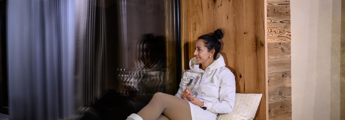 A woman is sitting relaxed on a cozy bench in front of a large window. She is holding a glass of water and enjoying the peaceful atmosphere. - The Peak Sölden - Sölden