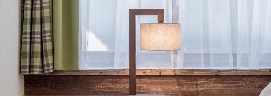A modern lamp stands on a wooden cabinet. The room has large windows with white curtains and a tranquil atmosphere. - The Peak Sölden - Sölden