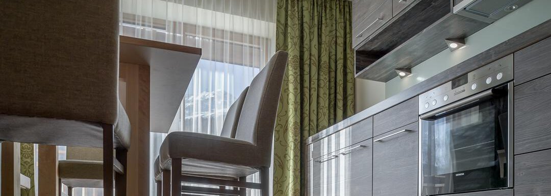 A modern kitchen with dark wooden cabinets and a built-in oven. The room has large windows with light curtains and a dining area with chairs. - The Peak Sölden - Sölden