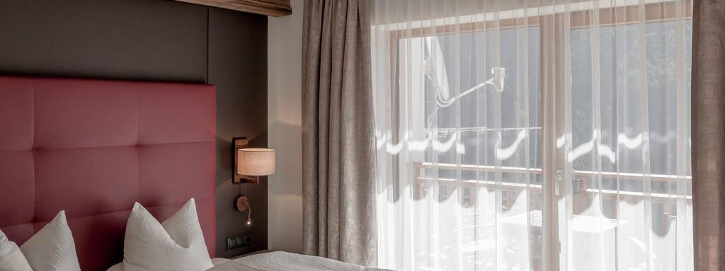 A modern bedroom with a red upholstered headboard and two single beds. Large windows let in plenty of light and offer a view of the balcony. - The Peak Sölden - Sölden