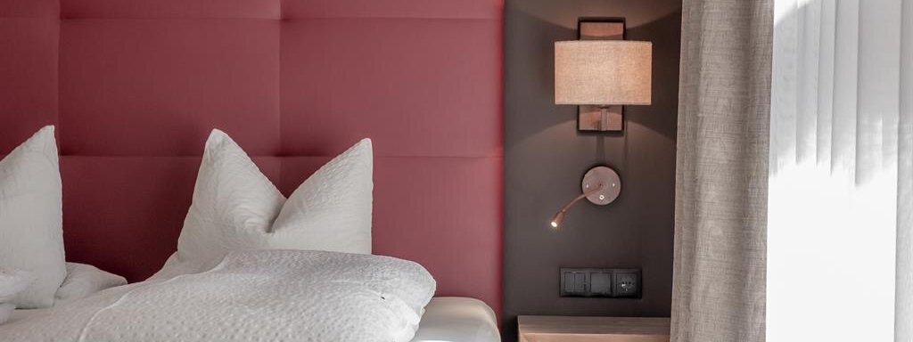 A modern bedroom with a red headboard and two large pillows. Next to the bed is a nightstand with a lamp. - The Peak Sölden - Sölden