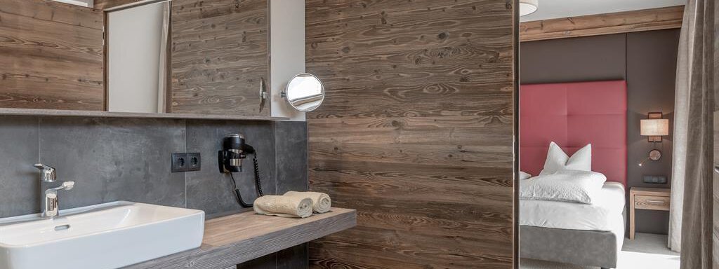A modern bathroom with a sink and wooden shelves. In the background, a cozy bedroom with a red headboard is visible. - The Peak Sölden - Sölden