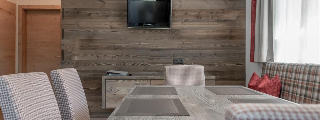 A modern dining area with a wooden table and striped chairs. A television hangs on the wall, and there are large windows with light curtains. - The Peak Sölden - Sölden