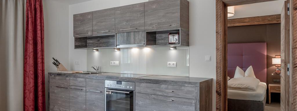 A modern kitchen with wooden cabinets and an integrated stove. In the background, a bedroom with a comfortable bed is visible. - The Peak Sölden - Sölden