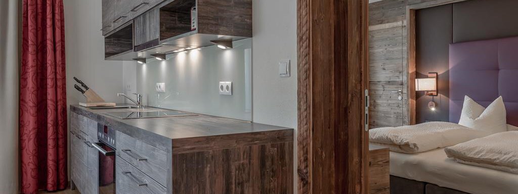 Modern kitchen with wooden cabinets and elegant lighting. In the background, there is a bedroom with a large bed visible. - The Peak Sölden - Sölden