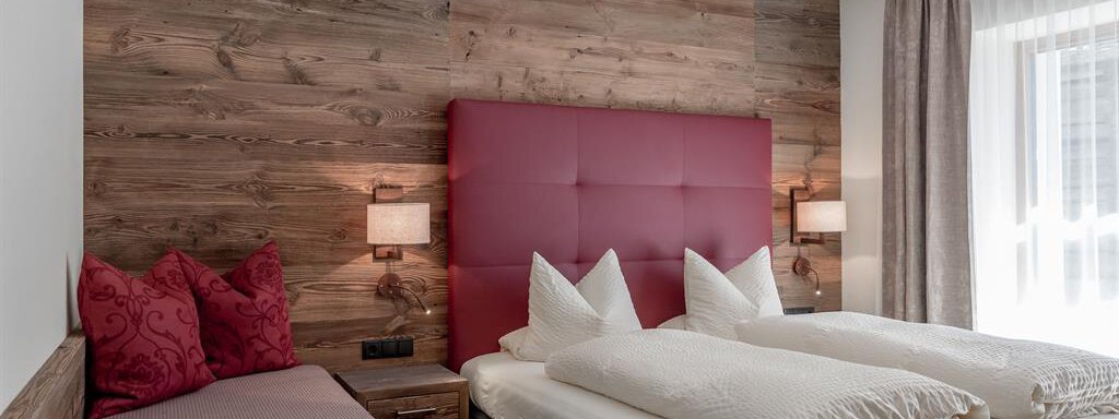 A stylish hotel room with a red headboard and light bed linens. The wood paneling and the windows create a cozy atmosphere. - The Peak Sölden - Sölden