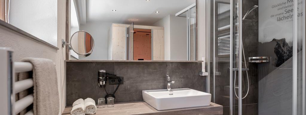 A modern bathroom with a shower and a large mirror. The wooden cabinet contains fresh towels in various colors. - The Peak Sölden - Sölden