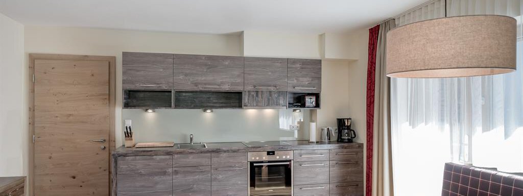 A modern kitchen with wooden cabinets and a cooking island. The dining area is bright and inviting, featuring a large table and a window with plenty of daylight. - The Peak Sölden - Sölden