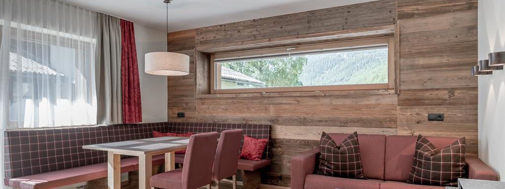 A cozy living space with wooden walls and large windows. There is a dining table, chairs, and a sofa in warm colors. - The Peak Sölden - Sölden